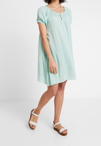 Robe mint verte à manches courtes avec un décolleté carré, des détails froncés et une coupe décontractée. Associée à des sandales blanches à semelles en bois.