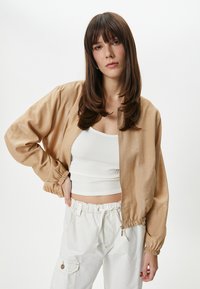 Beige, kevyt bomber-takki, jossa on kiiltävä pinta, joustavat hihansuut ja vetoketjusulku, yhdistetty valkoisen ribbiohuomen T-paidan ja cargo-housujen kanssa.