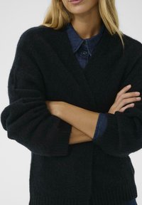 Schwarze, flauschige Strickjacke mit tiefem V-Ausschnitt, übergroßen Ärmeln und geripptem Saum, über einem dunklen Jeanshemd getragen.