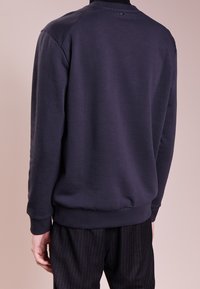 Uomo con una felpa a maniche lunghe di colore blu scuro e pantaloni neri a righe, mostrato di spalle su uno sfondo neutro.