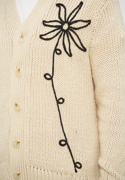 Μπεζ πλεκτή ζακέτα με μαύρη κεντητή λουλουδάτη διακόσμηση στην αριστερή πλευρά, με στρογγυλά κουμπιά μπροστά και υφασμένο σχέδιο.