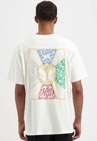 Crème T-shirt met korte mouwen met een kleurrafieke afbeelding op de achterkant. Het ontwerp omvat blauwe golven, groene en rode patronen, en een centraal embleem.