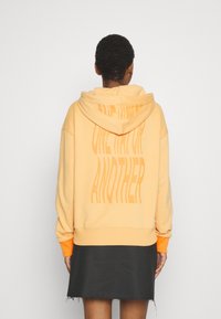 Felpa arancione con cappuccio, realizzata in un tessuto morbido e dal taglio oversize; presenta una grande grafica testuale tonale sul retro; abbinata a una gonna nera.