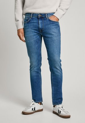 Jeans in denim blu chiaro con taglio slim, design a cinque tasche e cuciture a contrasto, abbinati a sneaker bianche con dettagli neri.