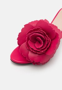 Chaussure à talon haut rose vif avec une grande décoration de fleur en cuir superposée sur la zone des orteils, sur fond blanc.
