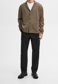 Homme portant un cardigan marron boutonné, un t-shirt blanc, un pantalon noir et des chaussures en cuir noir sur un fond blanc uni.