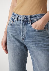 Jasnoniebieskie dżinsy o luźnym kroju, z delikatnym wyblaknięciem, miedzianymi nitami i szarym zapięciem na guzik. Widoczne kieszenie przednie.