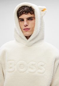 Hanorac din polar de culoare crem, cu glugă dotată cu urechi de urs și logo-ul "BOSS" embosat pe față. Textură moale și design confortabil.