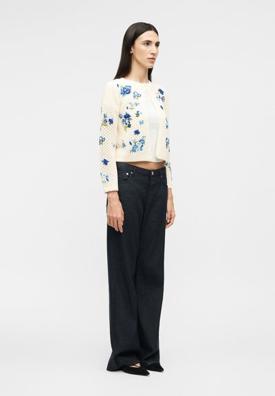 Γυναίκα με μακριά σκουρόχρωμα μαλλιά, φορώντας λευκό κοντό ζακέτα με μπλε ανθισμένη κεντητική, λευκό τοπ, φαρδιά μαύρη παντελόνα και μαύρα μυτερά παπούτσια.