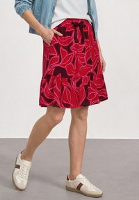 Femme portant une jupe à fleurs rouge et rose foncé avec une taille nouée, des baskets blanches et une veste bleu clair, debout avec une main dans la poche.