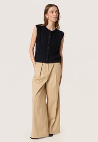Zwarte mouwloze ribgebreide cardigan met ronde hals, gecombineerd met beige wijde pantalon met voorplooien en hoge taille.