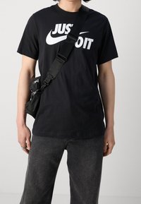 Person som bär en svart "Just Do It" Nike-t-shirt, mörkgrå jeans och en svart axelremsväska med dragkedjefickor.