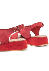Red suede wedge sandals with a wide strap and adjustable buckle, featuring an open heel and rounded toe, set on a tan platform sole.