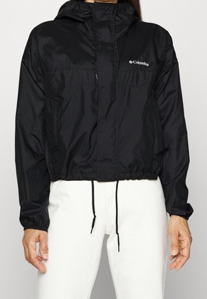 Person, die eine schwarze Columbia-Kapuzenjacke mit Kordelzug und weiße Hose trägt, von Hals bis Hüfte vor neutralem Hintergrund dargestellt.