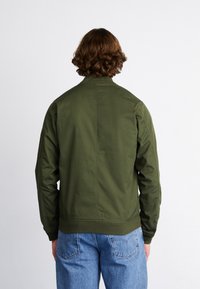 Veste aviateur vert olive avec un col côtelé et des poignets élastiques, fabriquée en tissu léger, associée à un jean bleu.