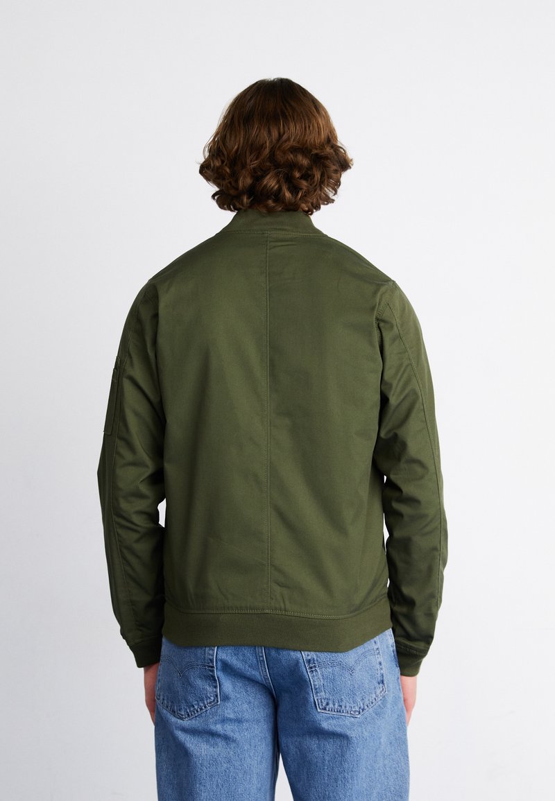 Veste aviateur vert olive avec un col côtelé et des poignets élastiques, fabriquée en tissu léger, associée à un jean bleu.