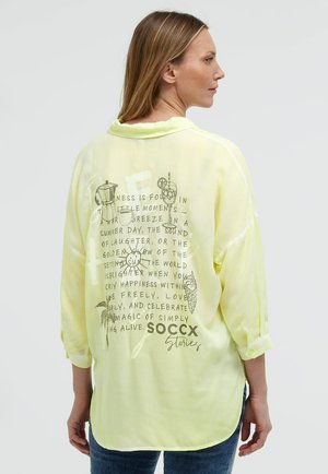 Frau trägt ein hellgelbes langärmeliges Shirt mit Text und kleinen Zeichnungen auf der Rückseite, kombiniert mit blauen Jeans.