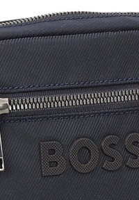 Tamsiai mėlynas nailono maišelis su tekstūruotu paviršiumi, turintis sidabrinę užtrauktuką ir iškilusį "BOSS" logotipą priekyje. Matomi subtilūs horizontalaus rašto elementai.