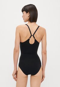 Μαύρο ολόσωμο μαγιό με ρυθμιζόμενους ιμάντες, κυκλικό άνοιγμα στην πλάτη και απαλή υφή υφάσματος. Μινιμαλιστικός σχεδιασμός, χωρίς σχέδια.