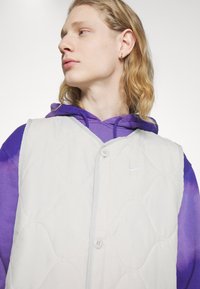 Hellgraune gesteppte Weste mit Knopfverschluss und lila Batikärmeln. Verfügt über das Nike-Logo auf der Brust und hat eine weiche Stofftextur.