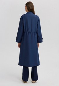 Tamnoplavi trench kaput s elastičnim strukom, dugim rukavima i dugmadima na manžetama; u kombinaciji s tamnoplavim hlačama. Glatka tekstura tkanine.