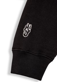 Manga de sudadera negra con puño acanalado y un logo de llama bordado en blanco cerca del codo, fabricada con tela suave.