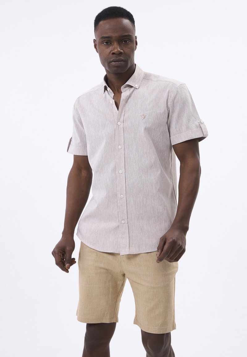 Uomo che indossa una camicia beige a maniche corte con bottoni e pantaloncini beige chiaro fino al ginocchio, in piedi davanti a uno sfondo bianco semplice.