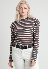 Langærmet stribet top med ribstrik i horisontale grønne, røde og hvide striber. Har flæser ved skuldrene og en rund halsudskæring, parret med højtaljede hvide bukser.