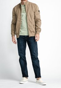 Man wearing beige jacket over light green graphic t-shirt, dark blue jeans rolled at the cuffs, and white slip-on shoes.