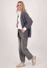 Grijze gebreide cardigan, witte shirt, grijze spijkerbroek en grijze sneakers. Zachte textuur, relaxte pasvorm, met geribbelde manchetten en een losse silhouet.