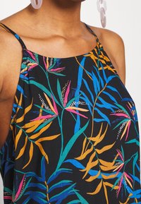 Nainen, jolla on päällä musta hihaton toppi, jossa on värikkäitä trooppisia lehtikuvioita, sekä läpikuultavat soikeat korvakorut, paljastaen hartiat ja kaulan.