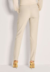 Beige slim-fit broek met een gladde textuur, voorzien van een zijvak en nadendetails. Gecombineerd met gouden hoge hakken sandalen.