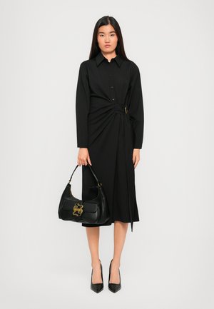 Femme portant une robe noire à manches longues avec une boucle latérale, tenant un sac à main noir avec des détails dorés, se tenant contre un fond uni.