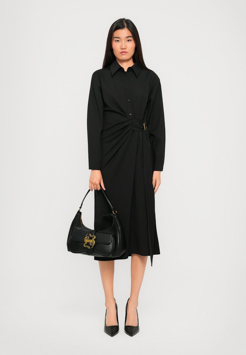 Woman wearing a long-sleeve black dress with a side buckle holding a black handbag with gold detailing, standing against a plain background.