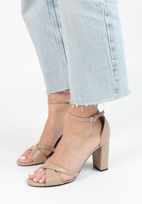 Beige hoge hakken sandalen met een enkelbandje, met gekruiste banden over de tenen en een blokhak. Gladde leren textuur.