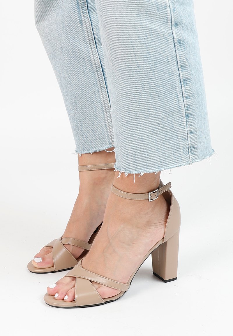 Beige hoge hakken sandalen met een enkelbandje, met gekruiste banden over de tenen en een blokhak. Gladde leren textuur.