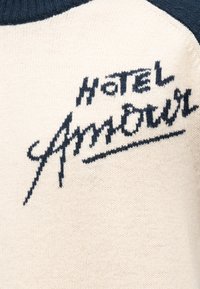 Κρεμ πλεκτό ύφασμα με μαύρο κείμενο "Hotel Amour" σε καλλιγραφικά και μπλοκ γράμματα σε κοντινή λήψη ενδύματος.