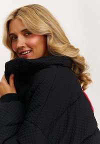 Blondýnka se usmívá, zvedá límec texturované černé prošívané bundy, s volnými kadeřemi na jednobarevném světle. 