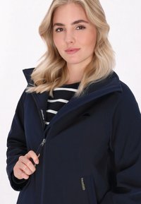 Žena s vlnitými blond vlasmi sa usmieva, oblečená v námorníckej bunde cez čierno-bielo pruhovanú košeľu, s rukami pri zipsu.