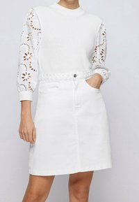 Kvinna iklädd en vit texturerad tröja med blommig eyelet-puffärmar och vit A-linjeformad kjol med flätad bälte, hand i fickan.