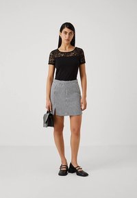 Musta pitsiga kaunistatud ülemine osa koos kõrge taljega musta-valge hundhammaste mustriga seelikuga, millel esiküljel lõhik; modell hoiab väikest musta kotti.