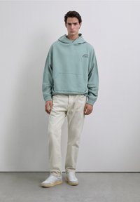 Sudadera con capucha azul claro con un bolsillo tipo canguro, combinada con pantalones blancos y zapatillas blancas con suelas color canela. Diseño sencillo con branding mínimo.