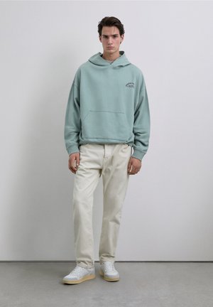 Lichtblauwe hoodie met een kangoeroezak, gecombineerd met witte broek en witte sneakers met beige zolen. Eenvoudig ontwerp met minimale branding.