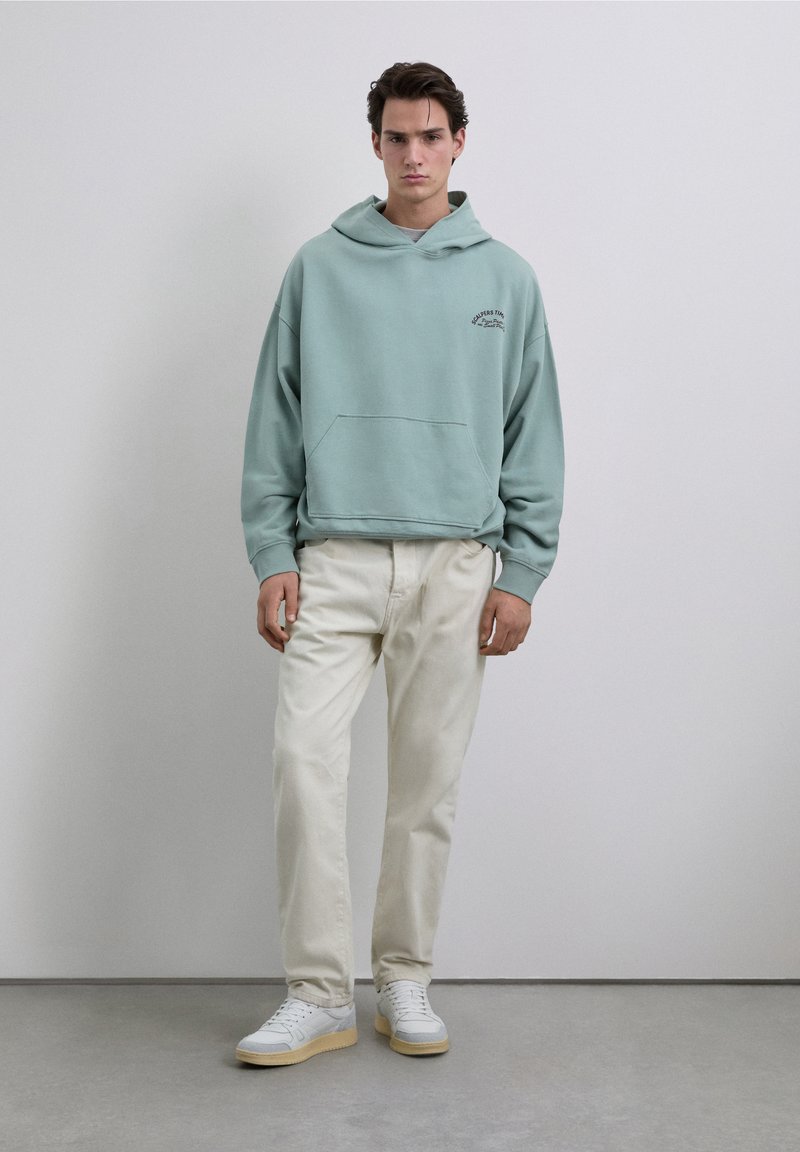 Sudadera con capucha azul claro con un bolsillo tipo canguro, combinada con pantalones blancos y zapatillas blancas con suelas color canela. Diseño sencillo con branding mínimo.