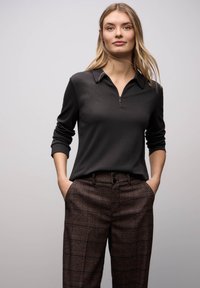 Schwarzes Polohemd mit Reißverschluss und langen Ärmeln, kombiniert mit braunen karierten Hosen, die zwei Knöpfe an der Taille und seitliche Taschen haben.