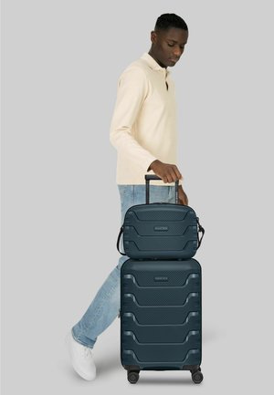 Man in beige lange mouwen shirt en spijkerbroek staand, houdt het handvat vast van twee gestapelde donkerblauwe harde koffers op wieltjes.