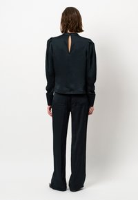 Schwarze Bluse mit Struktur und puffartigen Schultern sowie einem Schlüssellochdetail auf der Rückseite, kombiniert mit passenden geraden Hosen. Glatte Stoffoberfläche.