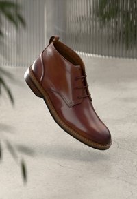 Scarpa da uomo in pelle marrone con lacci, suola in legno, sospesa sopra un pavimento in cemento testurizzato con piante sfocate in primo piano.