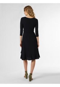 Schwarzes Kleid mit Dreiviertelärmel, taillierter Taille und schichtverklebtem Saum. Weicher Stoff, Reißverschluss hinten, getragen mit grünen Absätzen.