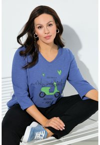 T-shirt a scollo a V blu con grafica di uno scooter verde e testo "Ciao Bella Rome 1987"; abbinata a pantaloni neri e sneaker azzurro chiaro.